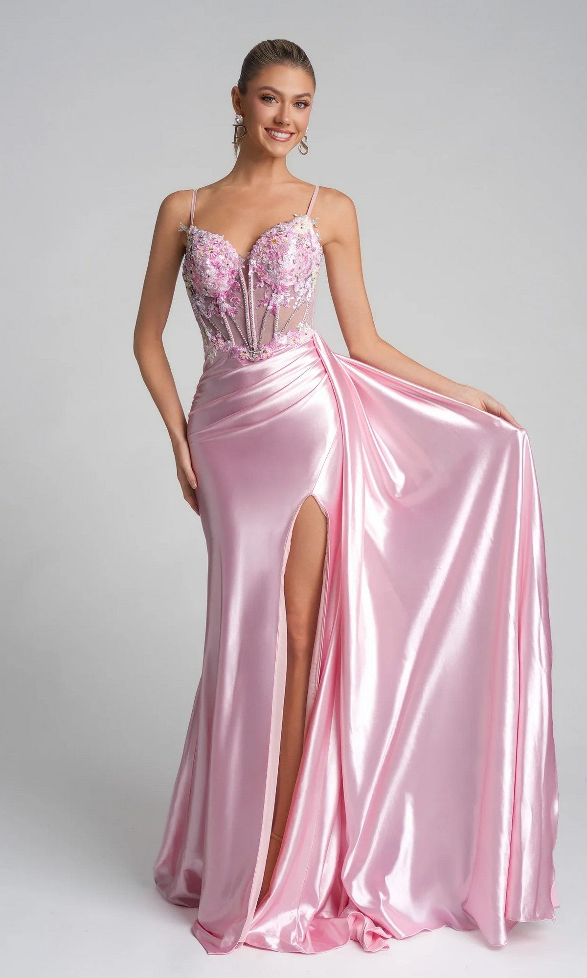 A woman wears the Portia and Scarlett PS26270 long pink prom dress with a shiny satin finish, thigh-high slit, embellished floral applique bodice, and spaghetti straps, holding out part of the skirt.